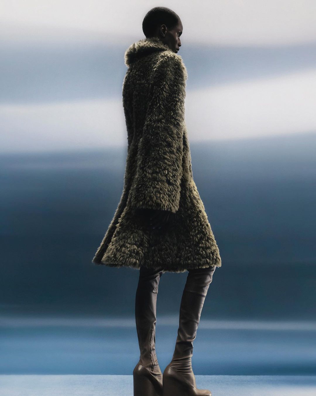 Fashion shot of a Black woman wearing a furry coat and thigh-high platform boots, posing against a soft blue background.