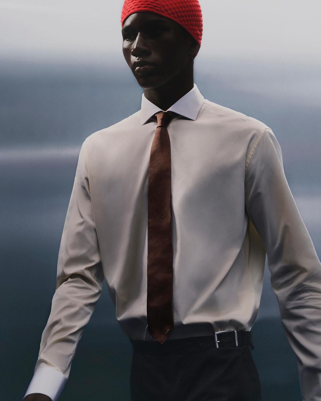Portrait of a Black male model in a button-down shirt, tie, and red knit cap against a blurred blue and grey background, fashion shoot.