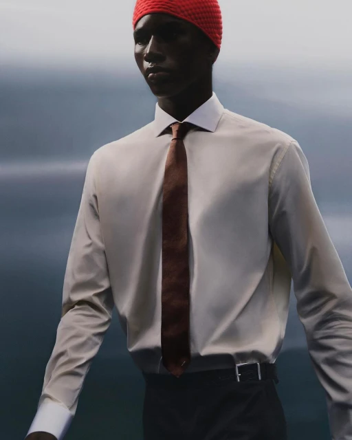 Portrait of a Black male model in a button-down shirt, tie, and red knit cap against a blurred blue and grey background, fashion shoot.