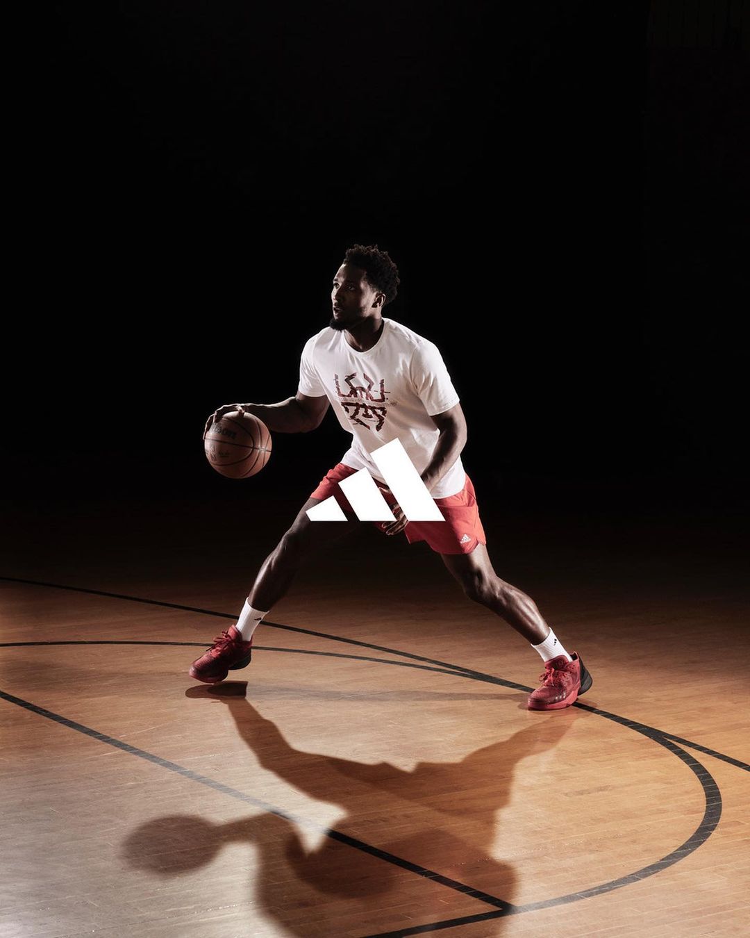 Athletic man dribbling basketball on court in a spotlight against a black background, wearing athletic apparel and basketball shoes.