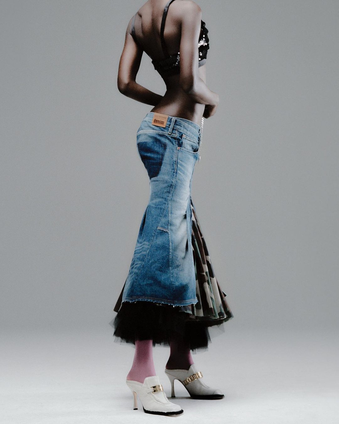 Woman in denim skirt and embellished top stands in a photo studio. The studio setting provides neutral backdrop highlighting the fashion elements.