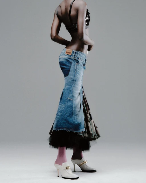 Woman in denim skirt and embellished top stands in a photo studio. The studio setting provides neutral backdrop highlighting the fashion elements.