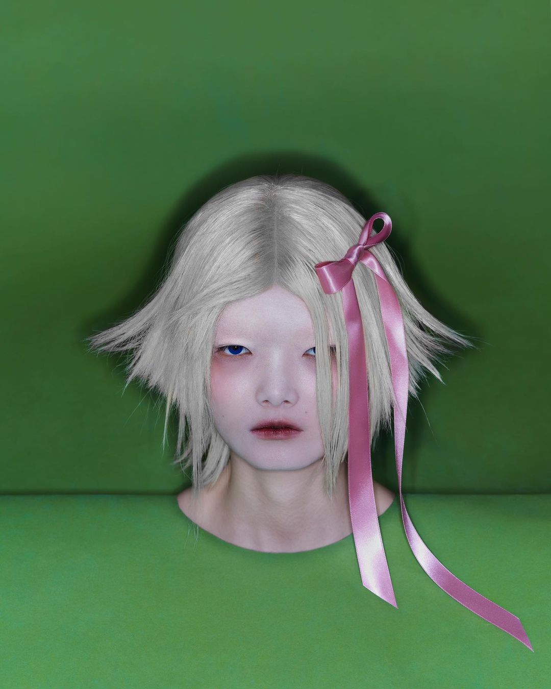 Close-up of a model with short white hair and a pink ribbon in her hair, posing against a green backdrop in a studio setting.