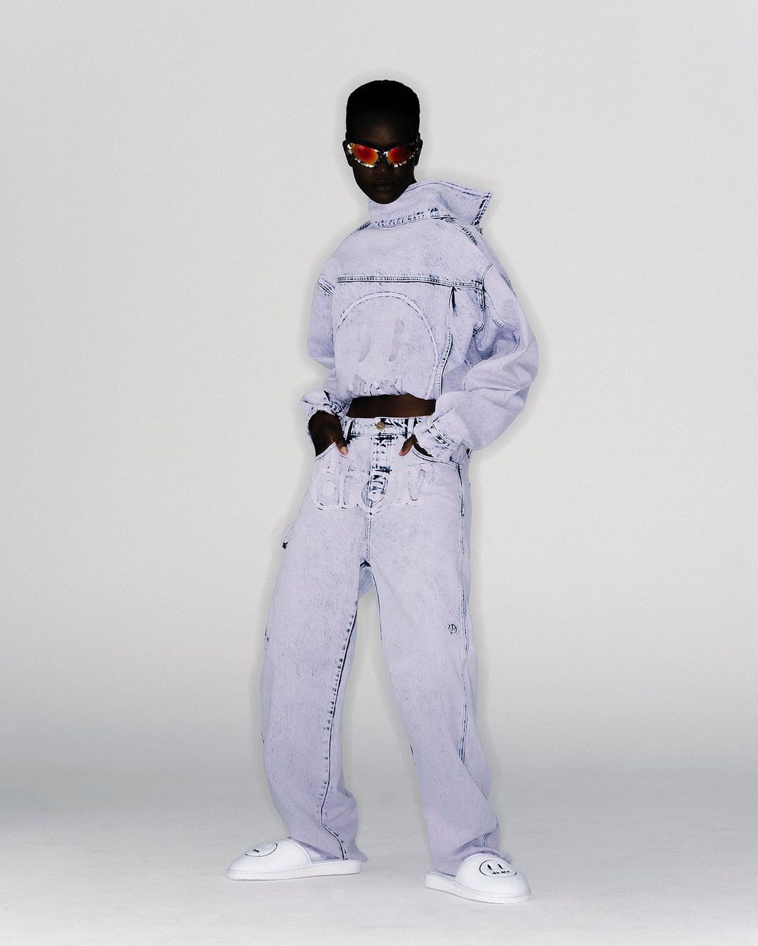 Adult female poses in an indoor studio wearing a matching light blue acid-wash denim jacket and baggy pants, along with white smiley-face slides.