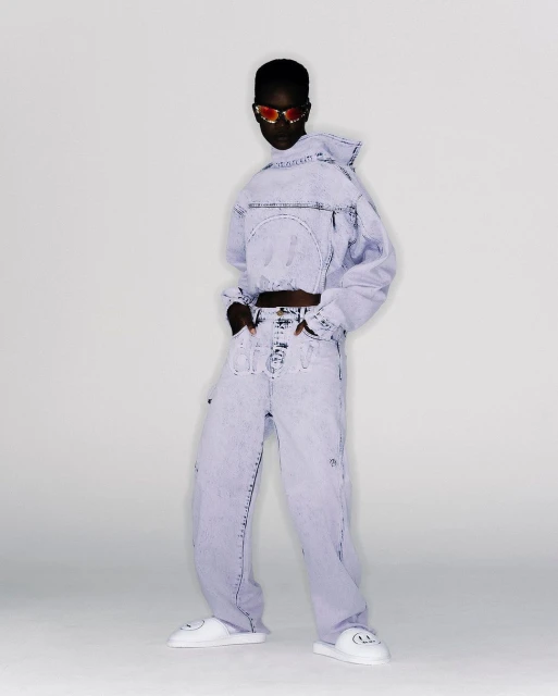 Adult female poses in an indoor studio wearing a matching light blue acid-wash denim jacket and baggy pants, along with white smiley-face slides.