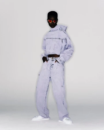 Adult female poses in an indoor studio wearing a matching light blue acid-wash denim jacket and baggy pants, along with white smiley-face slides.