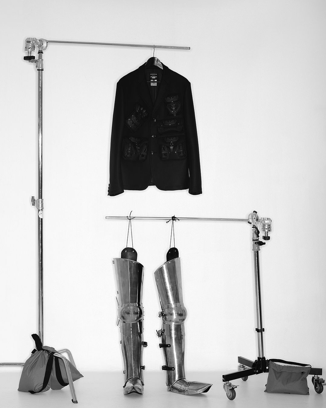 Black jacket hanging above metal armor leg coverings on metal racks in a photo studio with a white background, shot in black and white.