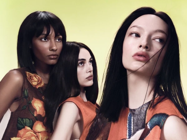 Three women pose in sleeveless dresses with floral and graphic prints against a bright yellow background, medium shot, fashion editorial.