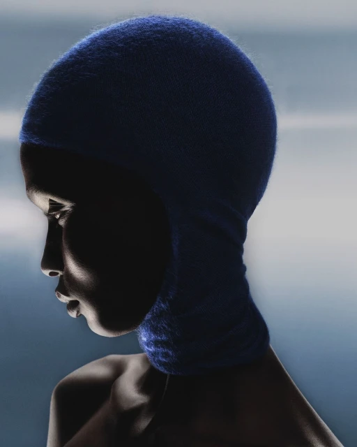 Profile shot of a dark-skinned woman wearing a blue knitted balaclava. The image features dramatic lighting and a soft blue gradient background.