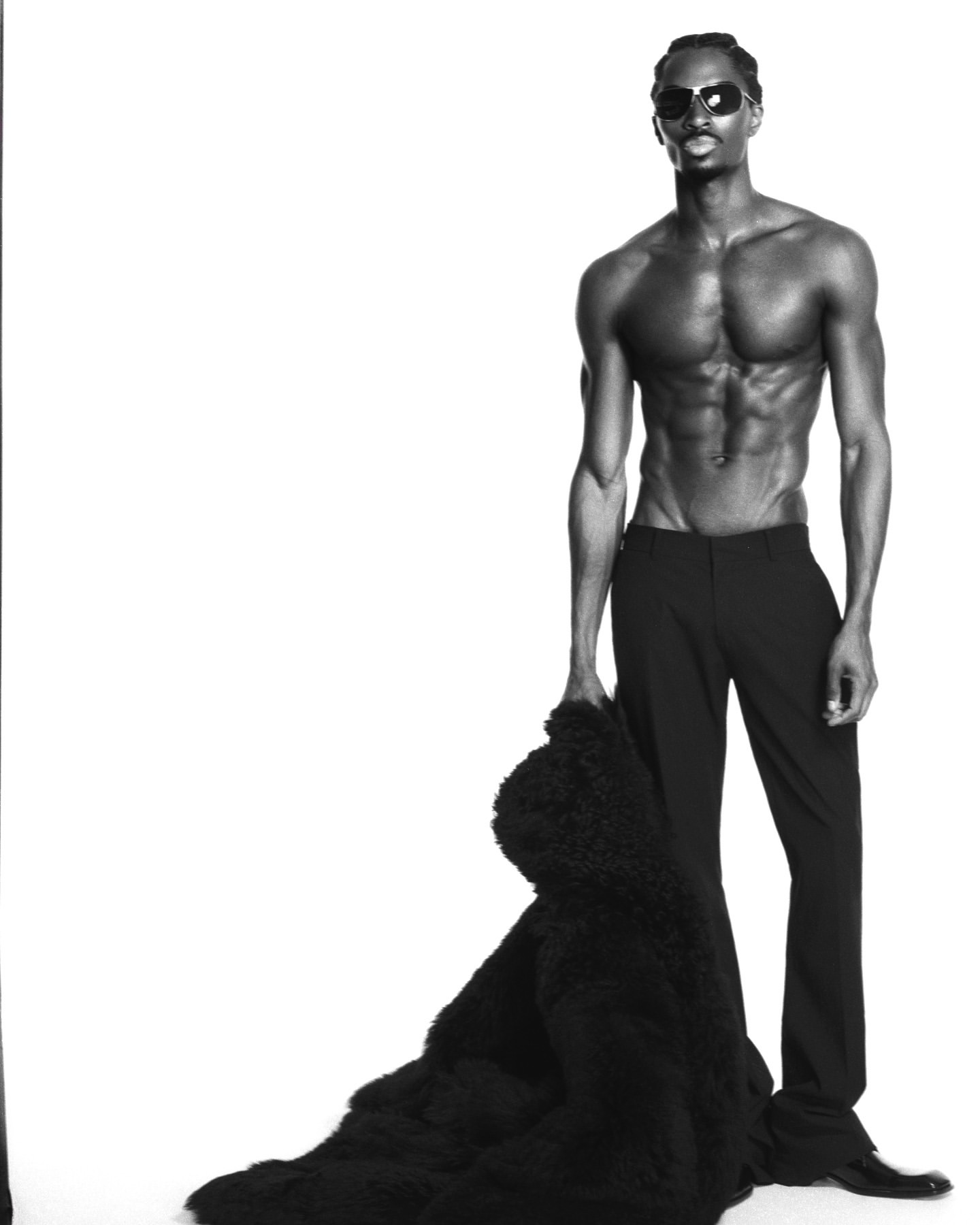 Black male fashion model poses shirtless in studio with sunglasses, wearing black trousers, holding a fur coat against a white backdrop.
