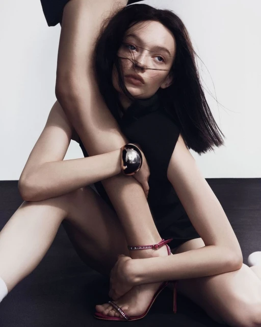 Fashion shot of a young adult woman in a sleeveless black shirt and pink high heels, posing on a black floor in a studio.