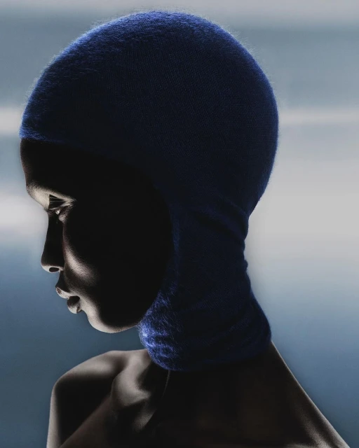 Profile of dark-skinned adult female in blue knit balaclava, illuminated by side lighting with diffused blue background.