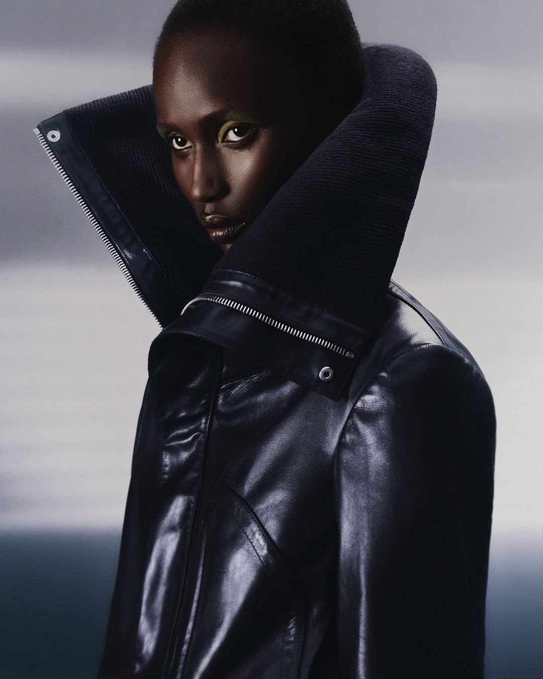 Fashion portrait of a black woman in a black leather jacket with a large collar against a gradient grey background in a studio setting.