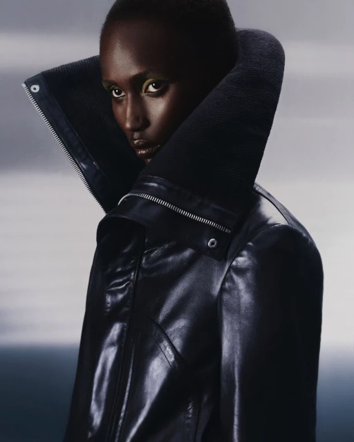 Fashion portrait of a black woman in a black leather jacket with a large collar against a gradient grey background in a studio setting.