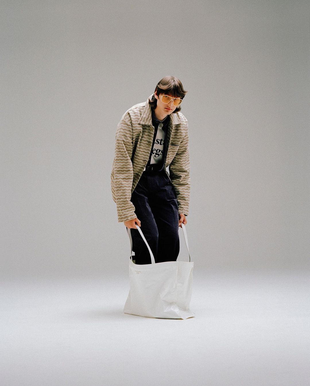 Full-length studio shot of a young adult wearing a striped jacket and yellow sunglasses, holding a white tote bag.