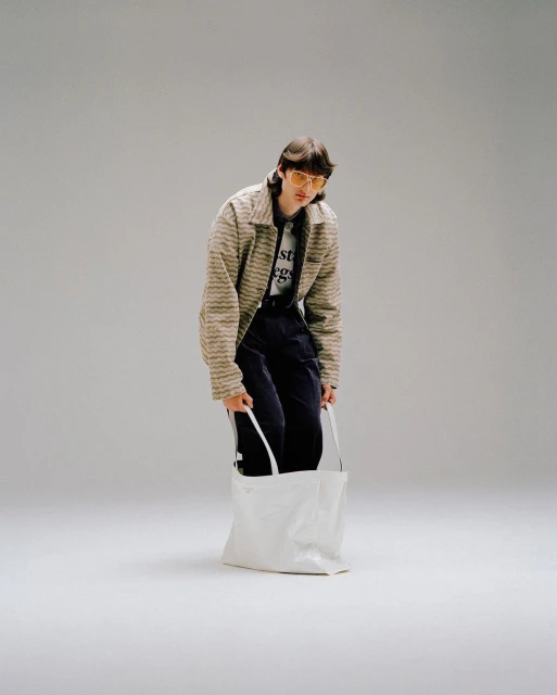 Full-length studio shot of a young adult wearing a striped jacket and yellow sunglasses, holding a white tote bag.