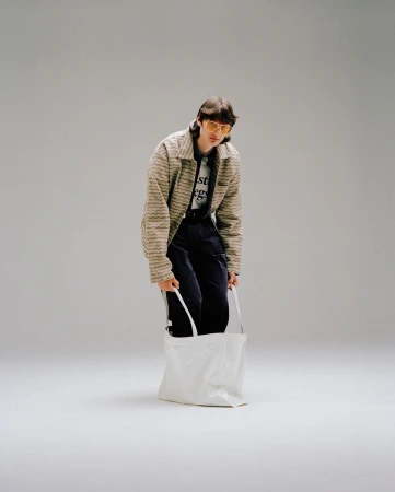 Full-length studio shot of a young adult wearing a striped jacket and yellow sunglasses, holding a white tote bag.