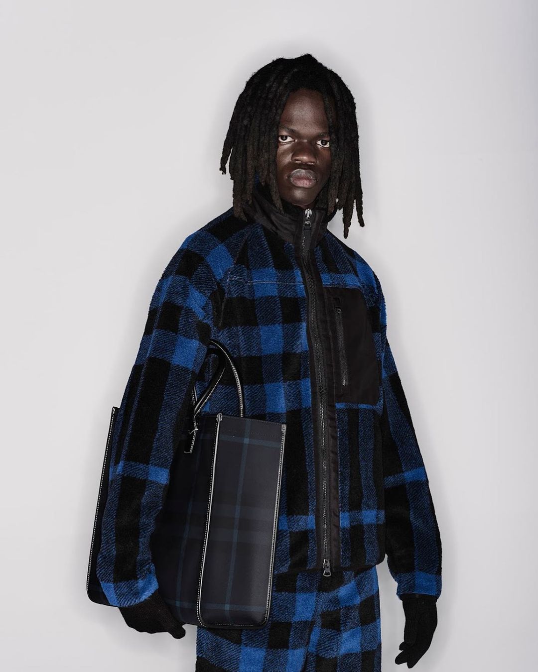 Model with dreadlocks wearing blue and black plaid fleece jacket holding structured tote bag against white background