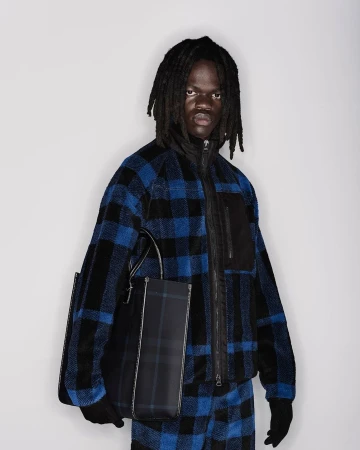 Model with dreadlocks wearing blue and black plaid fleece jacket holding structured tote bag against white background
