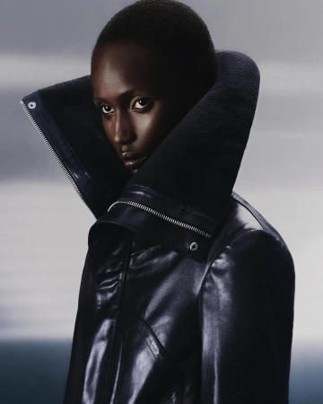 Dark-skinned woman with neutral expression in a black leather jacket with a large collar, studio lighting, against a light gray background.