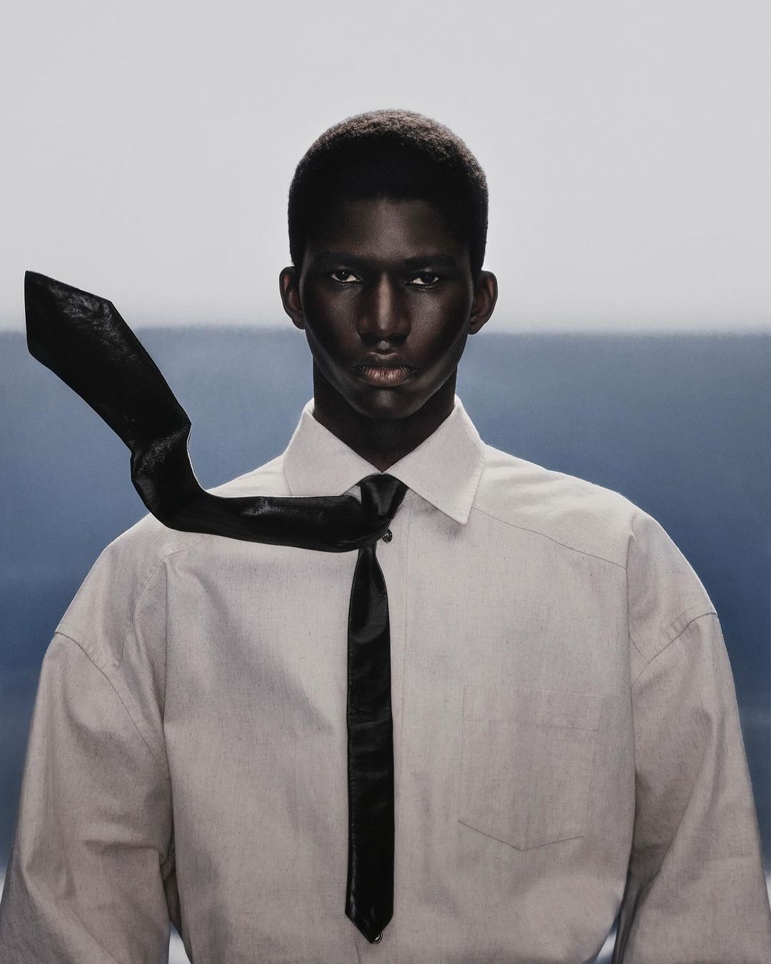 Studio shot of a black male model in a button-down shirt and tie against a split blue and white background, captured in a medium shot.