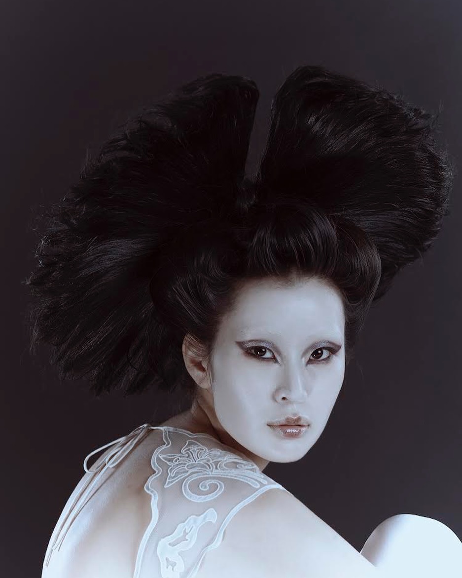 Portrait of Asian woman with white face makeup and sculptural black hairstyle against a dark backdrop in a beauty or fashion context.