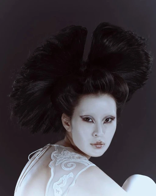 Portrait of Asian woman with white face makeup and sculptural black hairstyle against a dark backdrop in a beauty or fashion context.