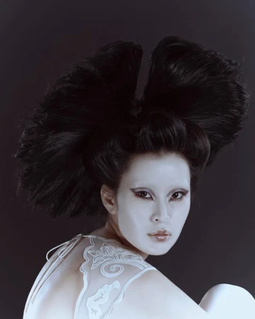 Portrait of Asian woman with white face makeup and sculptural black hairstyle against a dark backdrop in a beauty or fashion context.