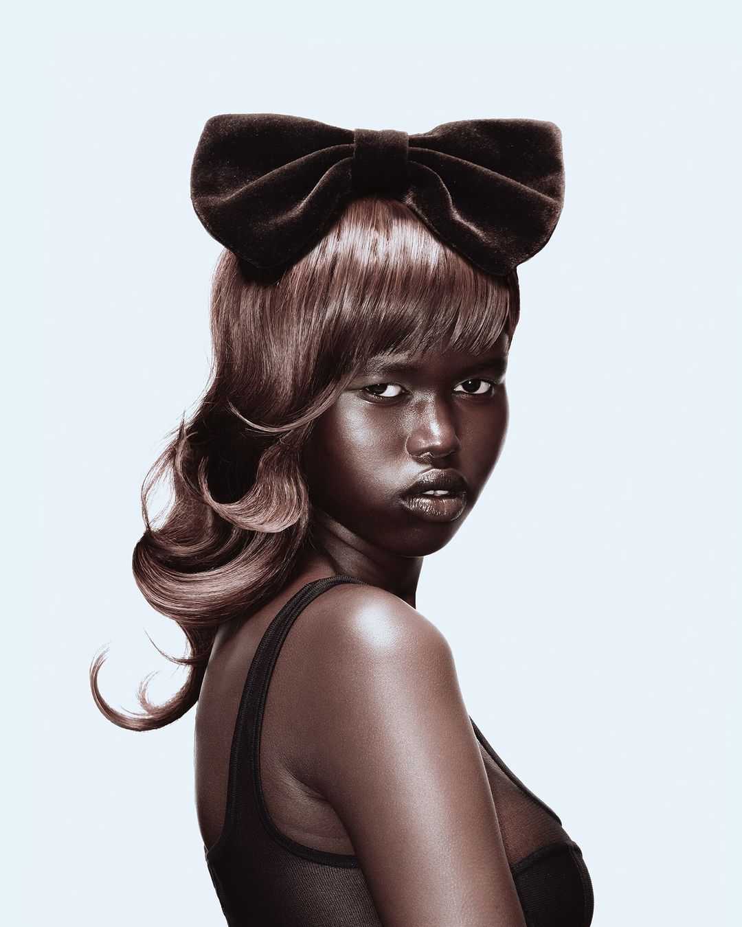 Close-up portrait of a woman with dark skin and a large black bow on her head, wearing a dark top against a white background.