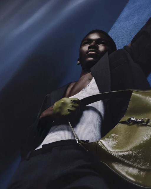 Fashion shot of an adult male model wearing a black blazer, white tank top, black pants, and holding an olive green purse.