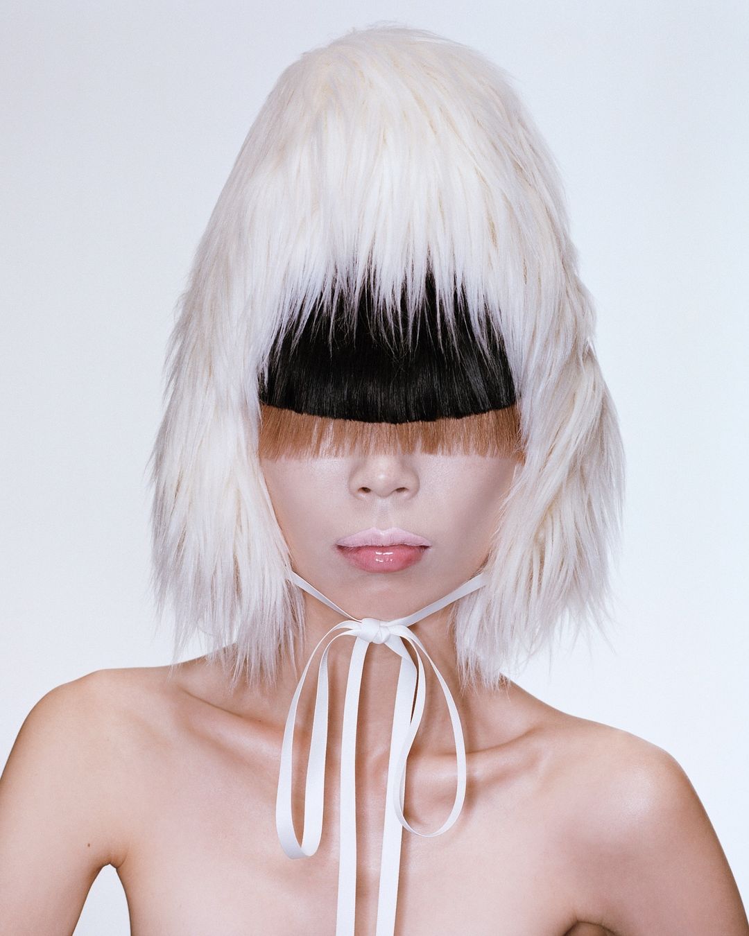 Striking studio portrait of a person with fair skin wearing a unique white fur hat with black and brown fringe covering their eyes.
