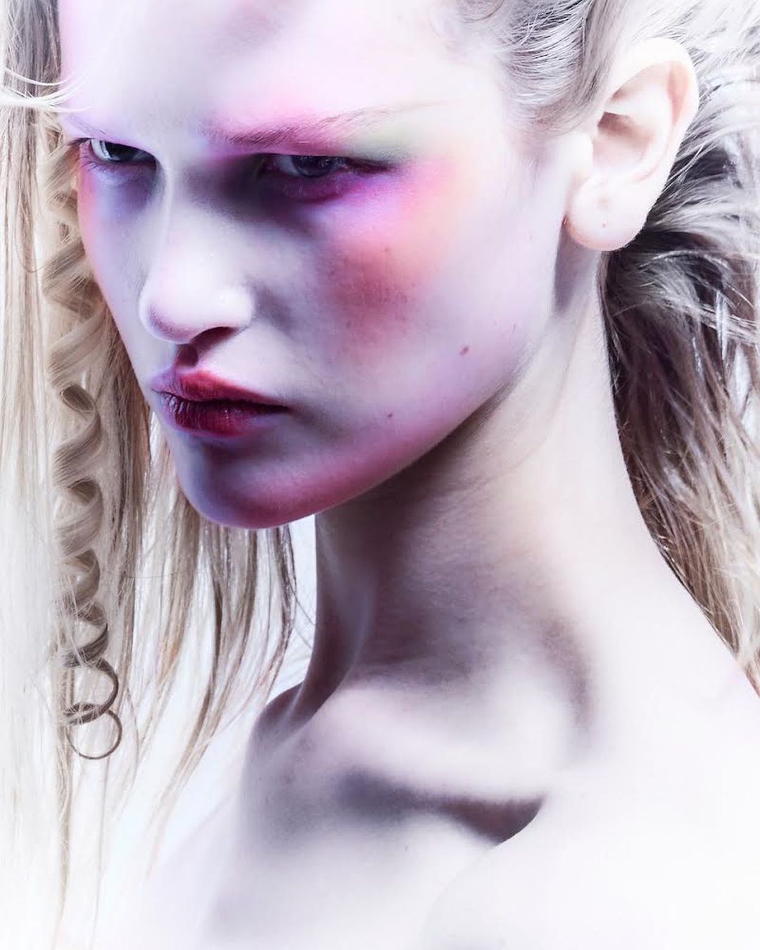 Close-up portrait of a female model with creative makeup in pink, purple, and green tones, set against a bright, neutral background.