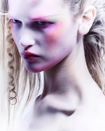 Close-up portrait of a female model with creative makeup in pink, purple, and green tones, set against a bright, neutral background.