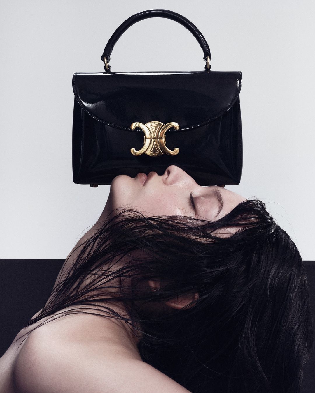 Close-up portrait of a woman with dark hair balanced beneath a black patent leather handbag featuring a gold clasp.