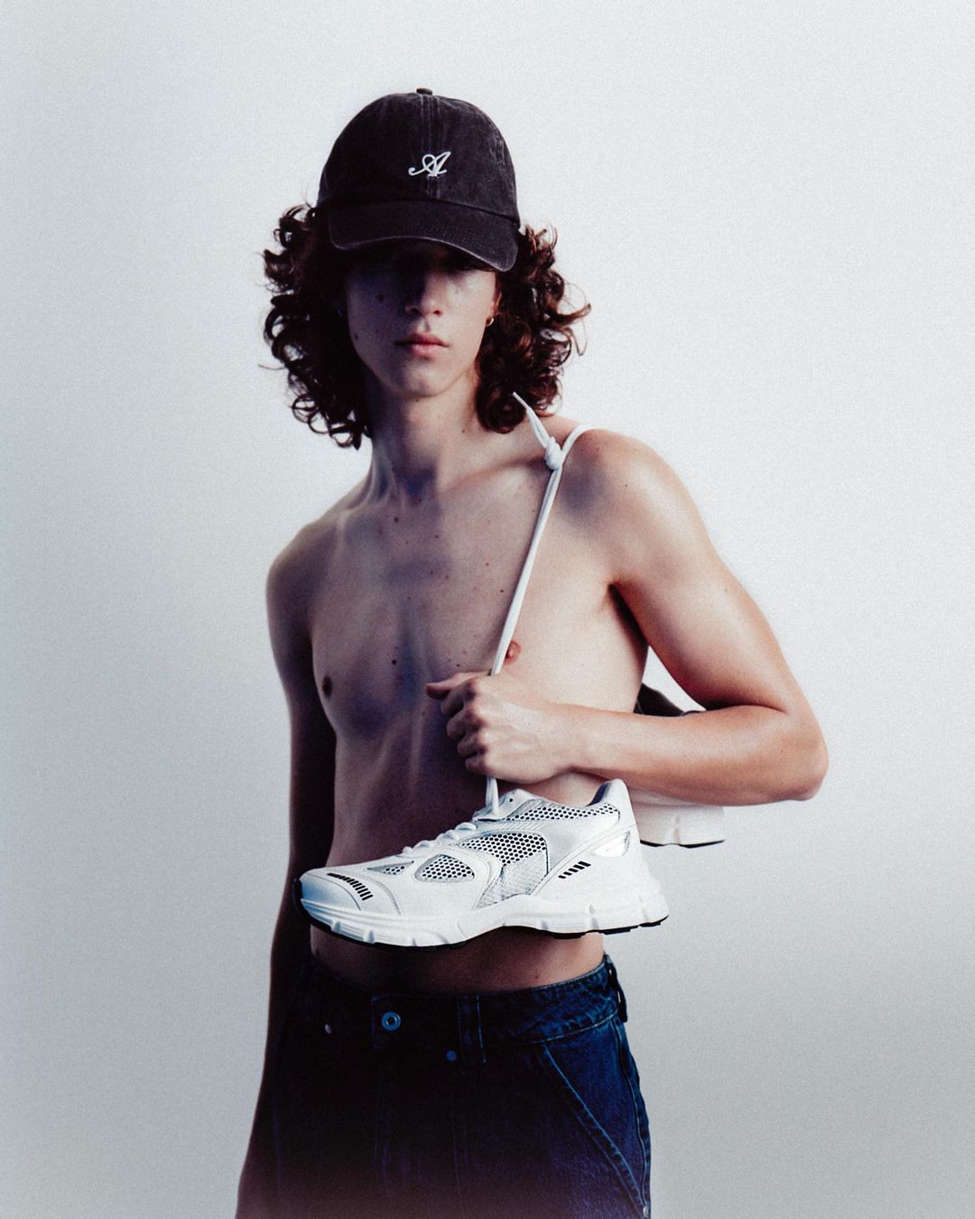 Young adult male with curly hair wearing a dark baseball cap and jeans, carrying a pair of white athletic shoes on his shoulder.