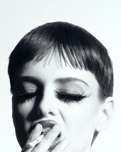Black and white portrait of a woman with exaggerated eyelashes smoking a cigarette, lips slightly parted and eyes closed.