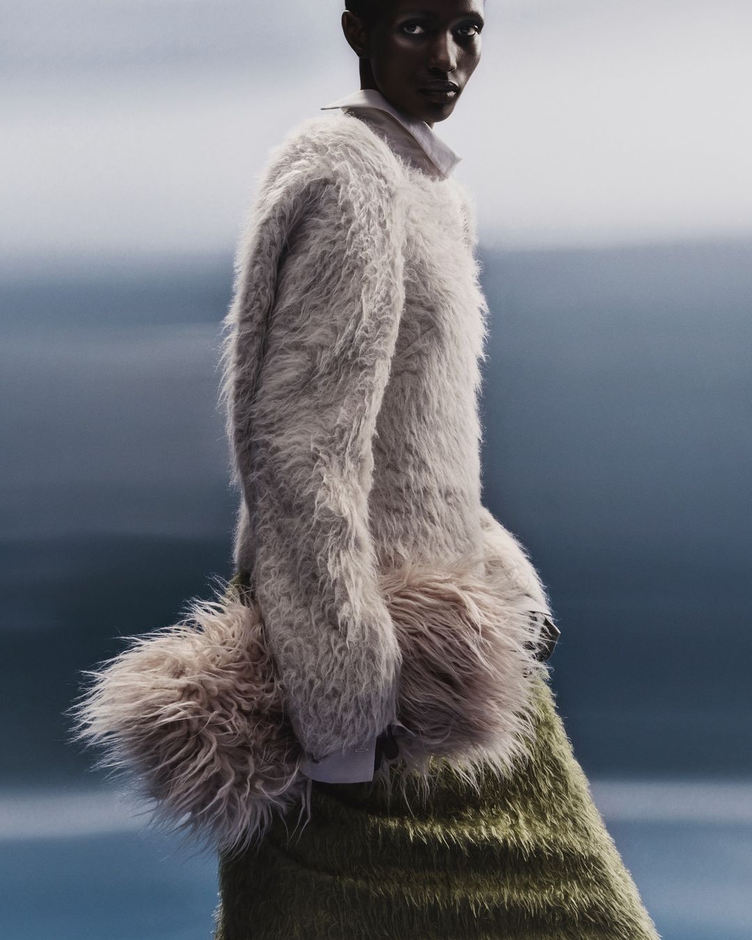 Fashion shot of Black woman in a furry sweater and olive skirt, posing against an out-of-focus gray and blue gradient backdrop.