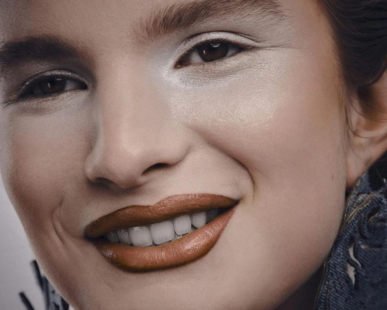 Close-up of a smiling woman with light skin, wearing a denim jacket, and light eyeshadow and lipstick in a brightly lit indoor photo.