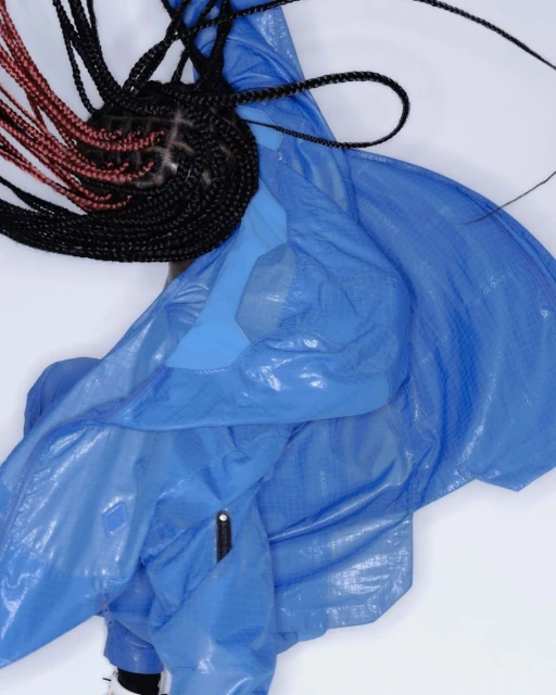 Overhead shot of a person with black and red braids wearing a shiny blue hooded jacket against a white backdrop in a fashion setting.