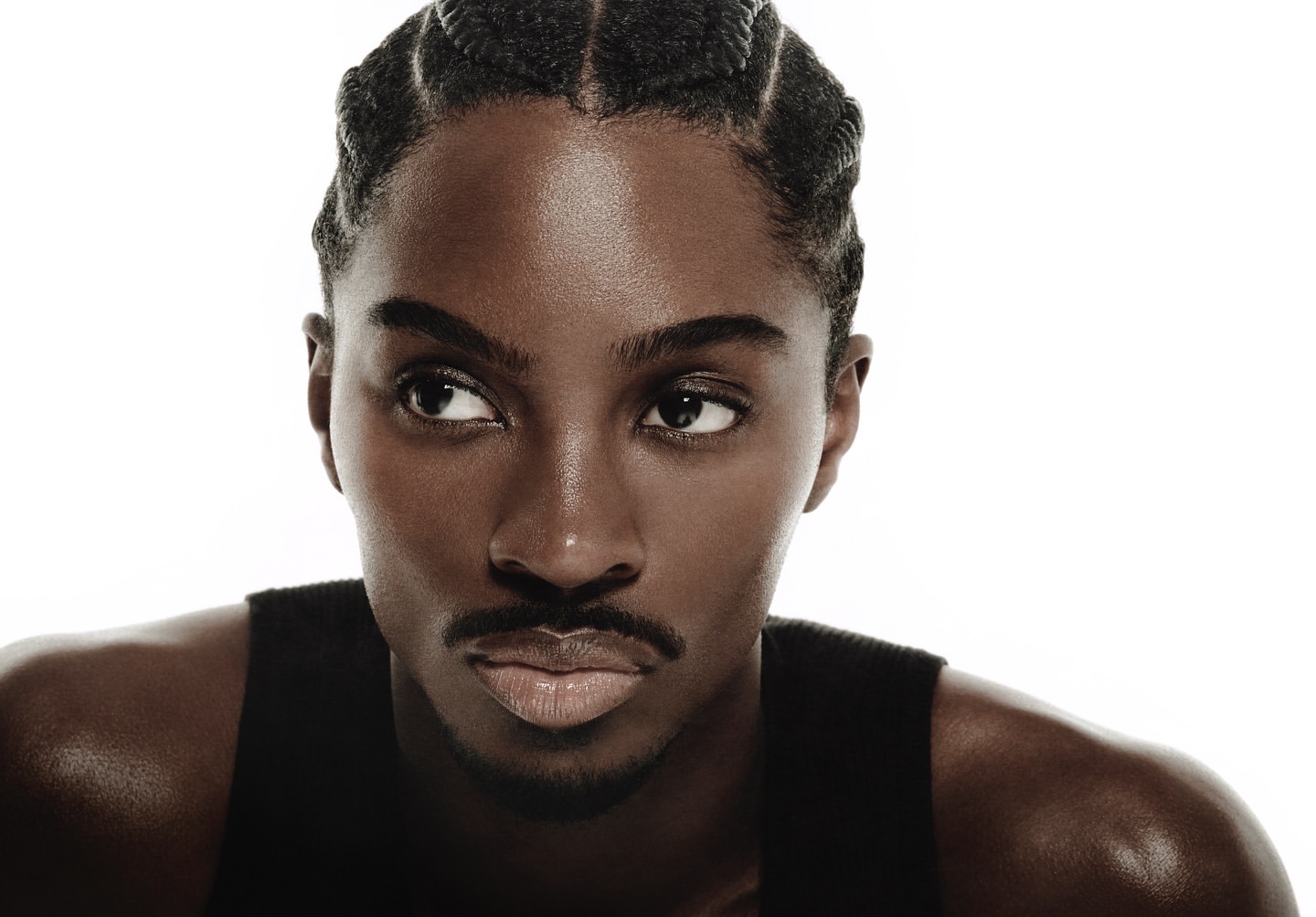 Close-up portrait of an adult male with dark skin and cornrow braids looking off to the side, wearing a black tank top against a white background.