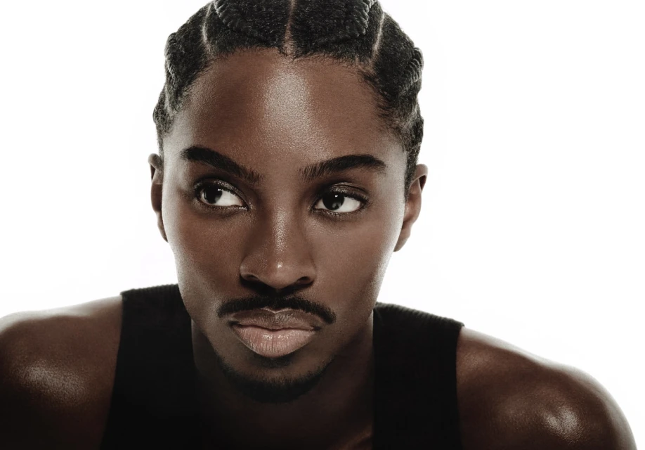 Close-up portrait of an adult male with dark skin and cornrow braids looking off to the side, wearing a black tank top against a white background.