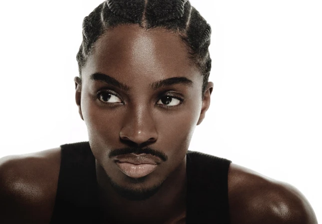 Close-up portrait of an adult male with dark skin and cornrow braids looking off to the side, wearing a black tank top against a white background.