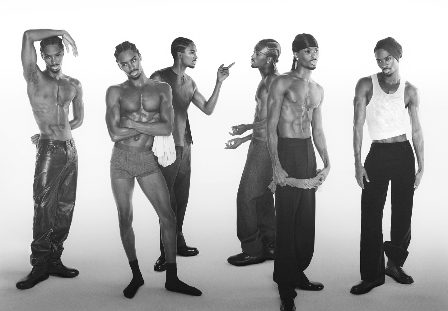Group of six young Black men in a photo studio wearing casual clothing, with bare chests, leather pants, and a white background.