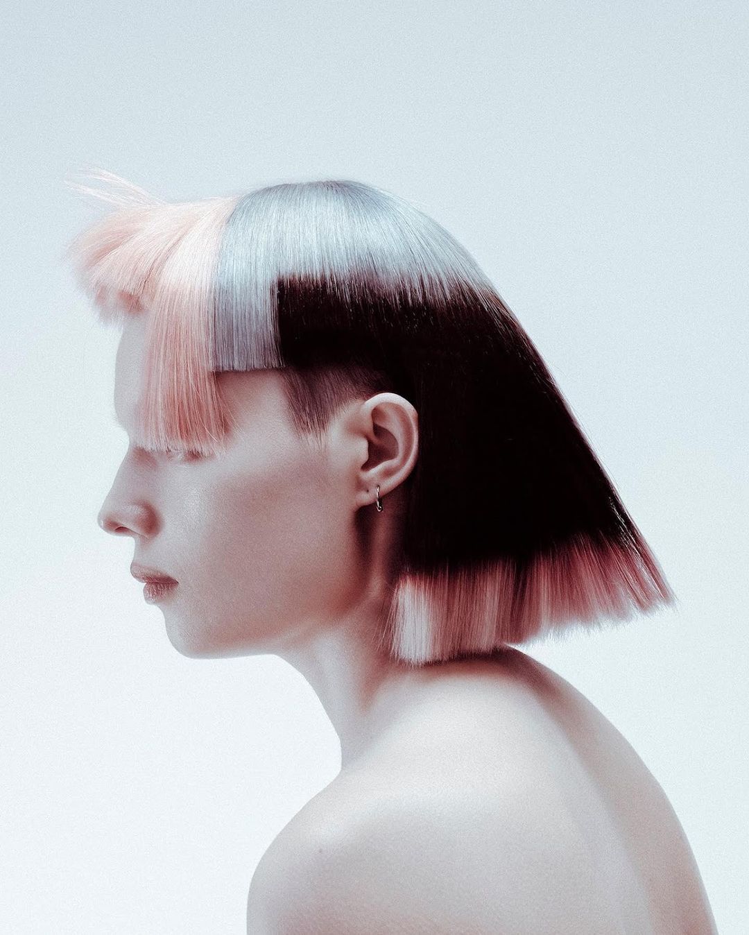 Profile shot of a person with an asymmetrical blunt bob haircut. The hair is colored in shades of black and pink, against a light backdrop.