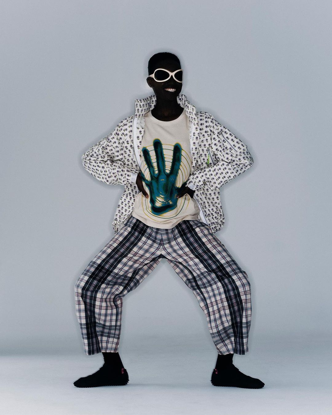 Person with dark skin poses in a graphic tee, jacket and plaid pants in a studio with a neutral gray backdrop, smiling in white glasses.