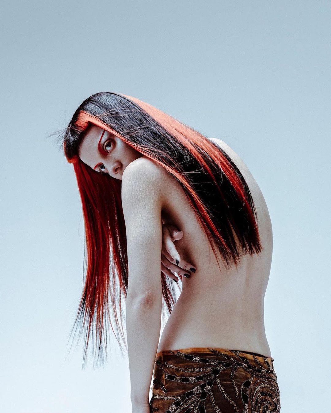 An adult woman with dark and red hair looks back over her shoulder in a studio shot. She's partially nude with a patterned skirt.