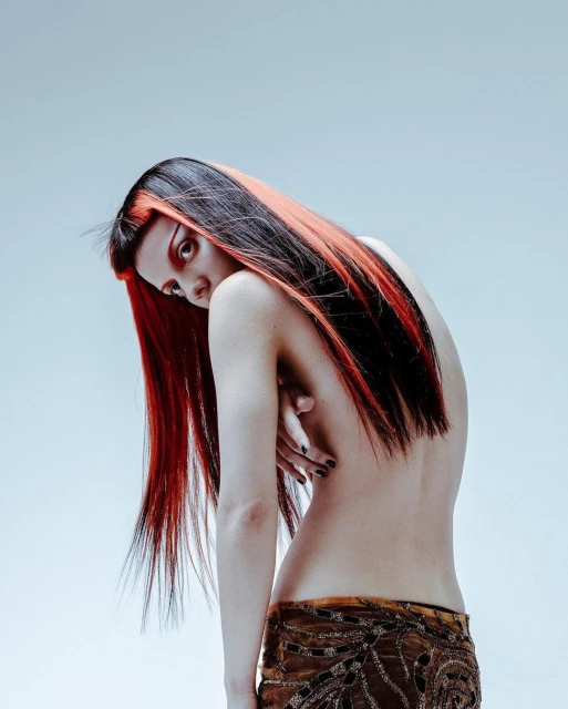 An adult woman with dark and red hair looks back over her shoulder in a studio shot. She's partially nude with a patterned skirt.