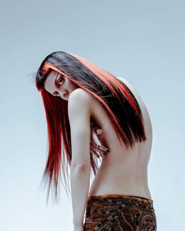 An adult woman with dark and red hair looks back over her shoulder in a studio shot. She's partially nude with a patterned skirt.