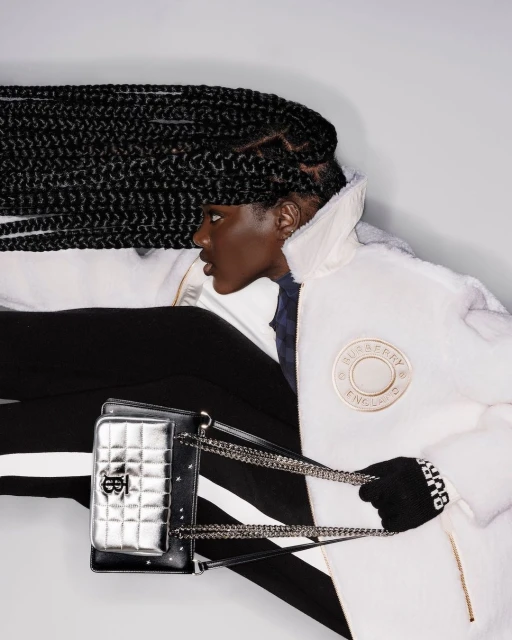 Fashionable woman with long braids wears a white coat and black striped pants while holding a silver purse in a photo studio.