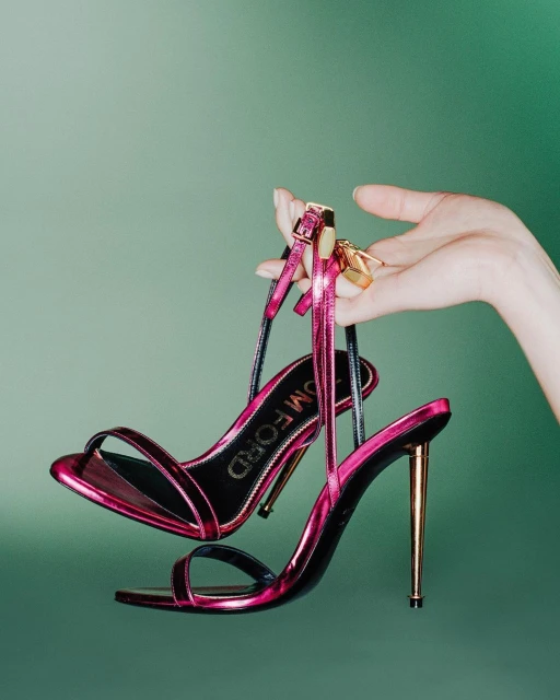 Pink Tom Ford stiletto sandal is held by a hand against a green gradient background in a product shot. Fashion and accessories.
